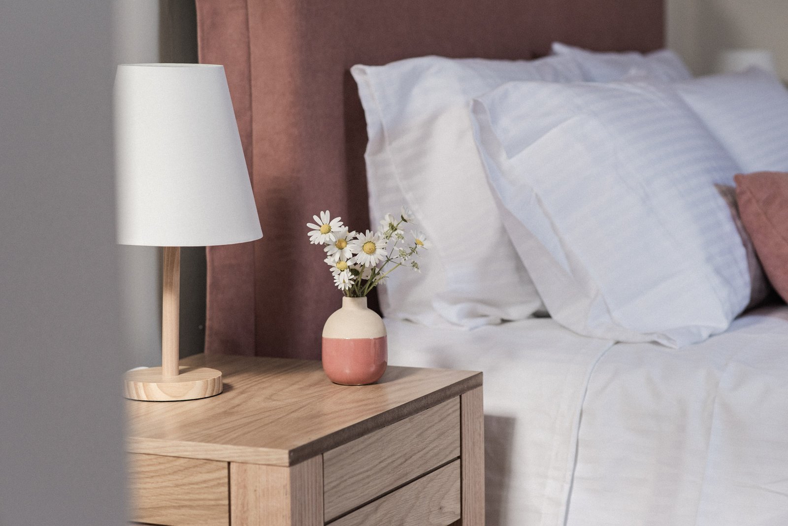 Bed with pale pink linens and bedside table decorated with a vase of fresh daisies